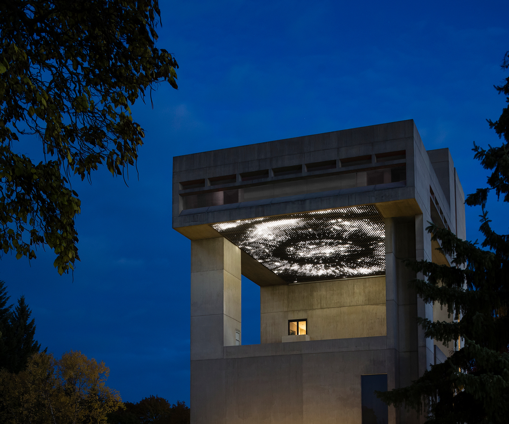 A pattern of lights appears beneath the cantilevered roof of a concrete building at night with tall trees on either side