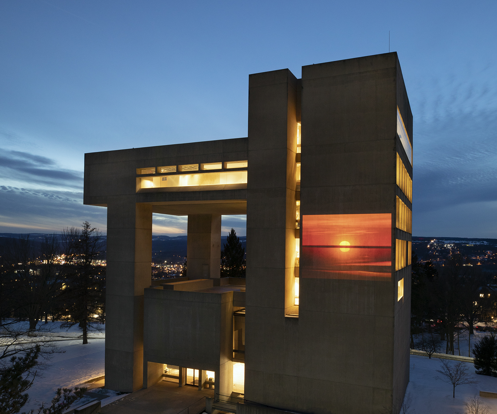 A yellow and orange image of the horizon and sun appears on the facade of the Johnson Museum building at twilight in the snow