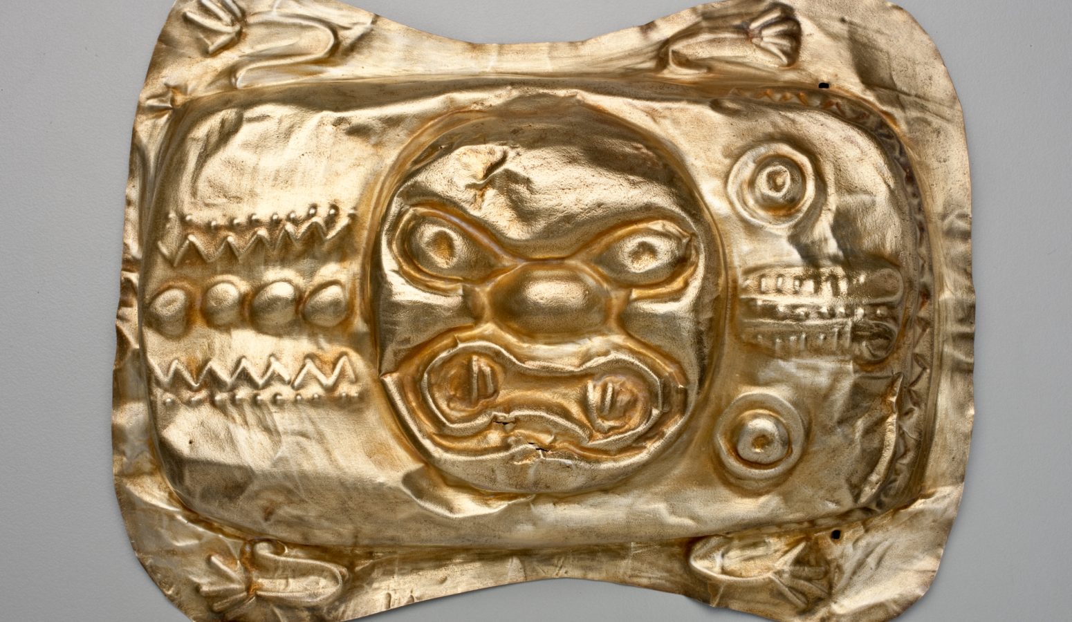 A gold metal object with embossed designs around a snarling animal face