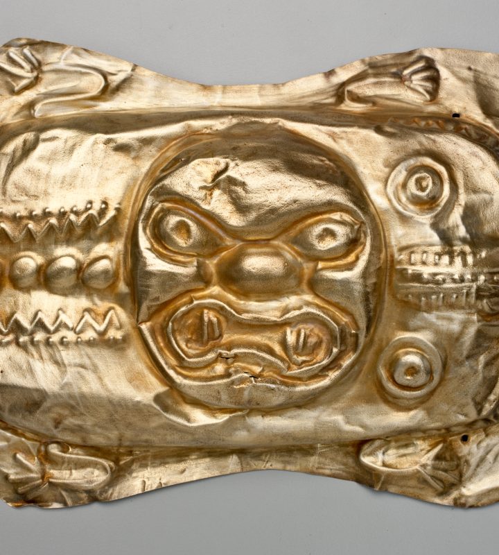 A gold metal object with embossed designs around a snarling animal face