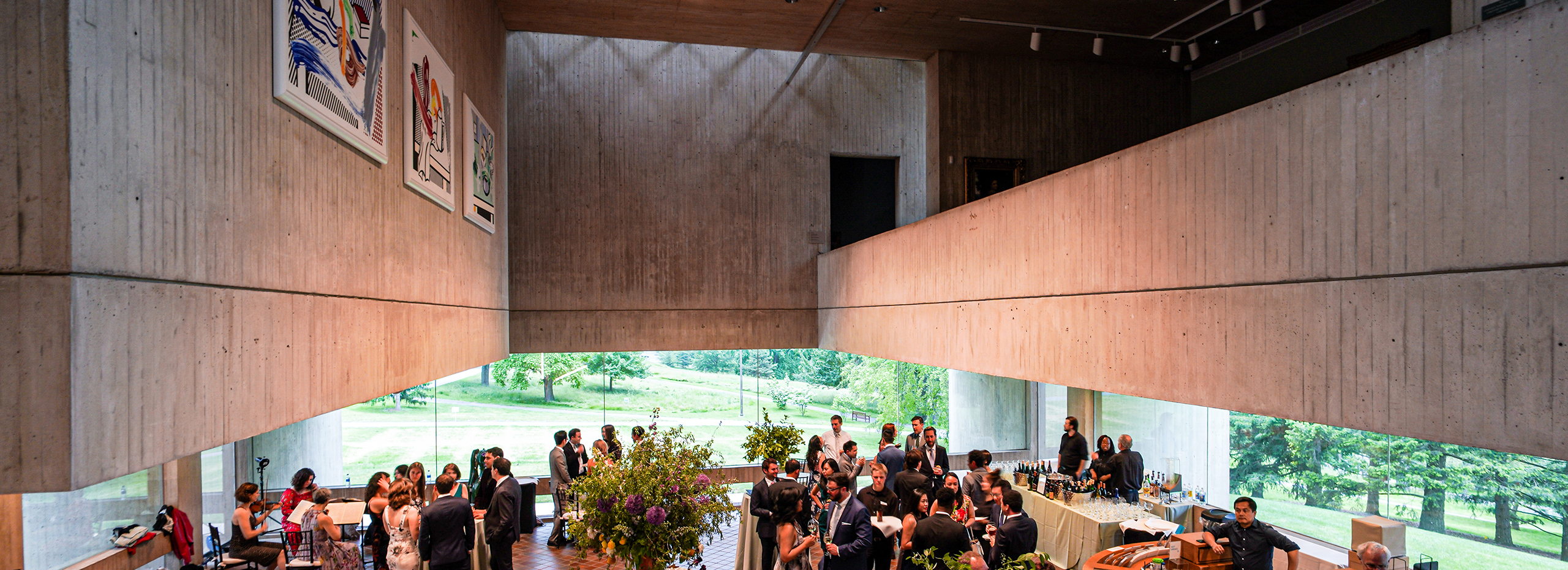 People gather in a large lobby with wide windows, concrete walls, and large paintings