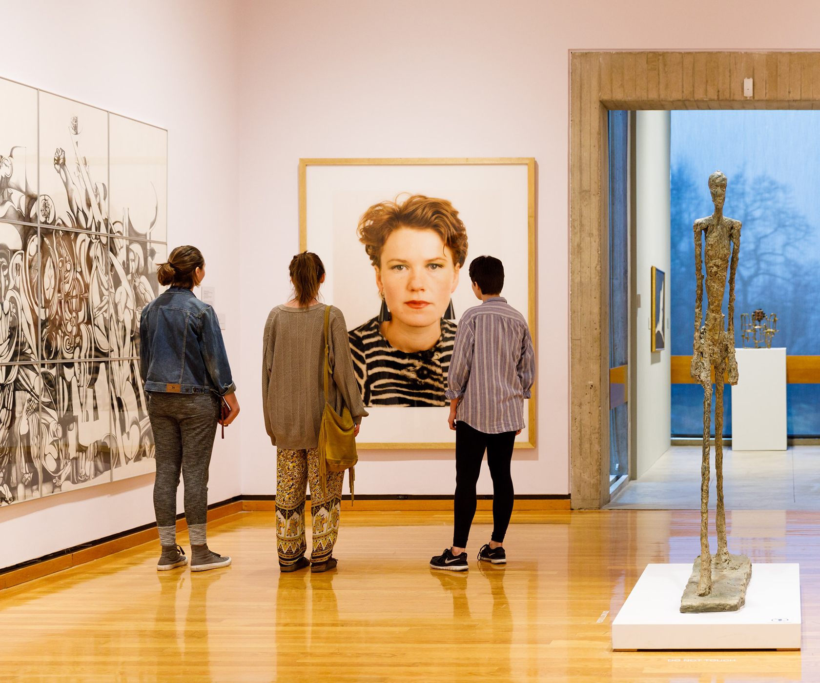 Three people look at a large photographic portrait of a white woman with short hair in a museum gallery with sculpture and other art on the light pink walls