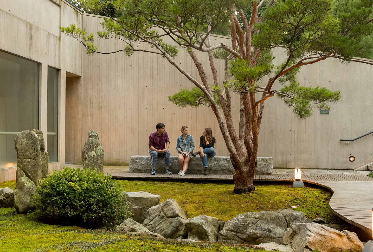 Three people sit on a stone bench overlooking a Japanese garden with moss, stones, and a large tree