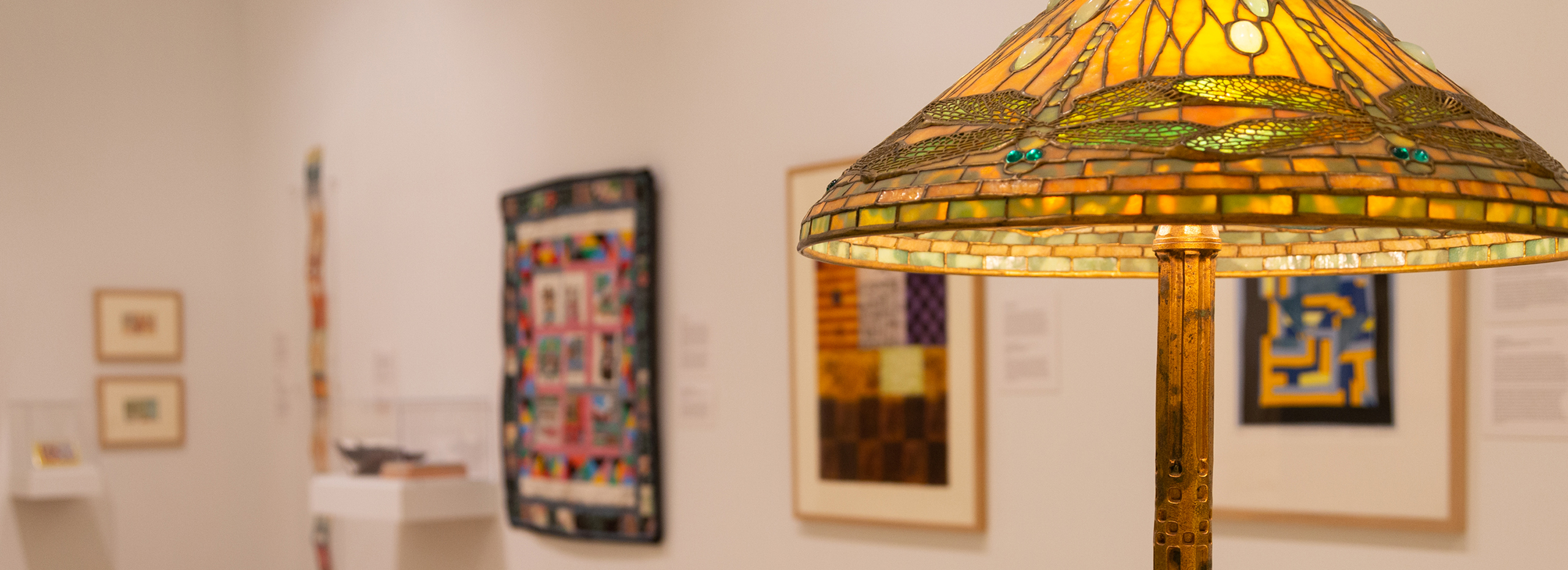 A lit stained glass lamp is in the foreground at right and a variety of colorful artworks are out of focus in the background