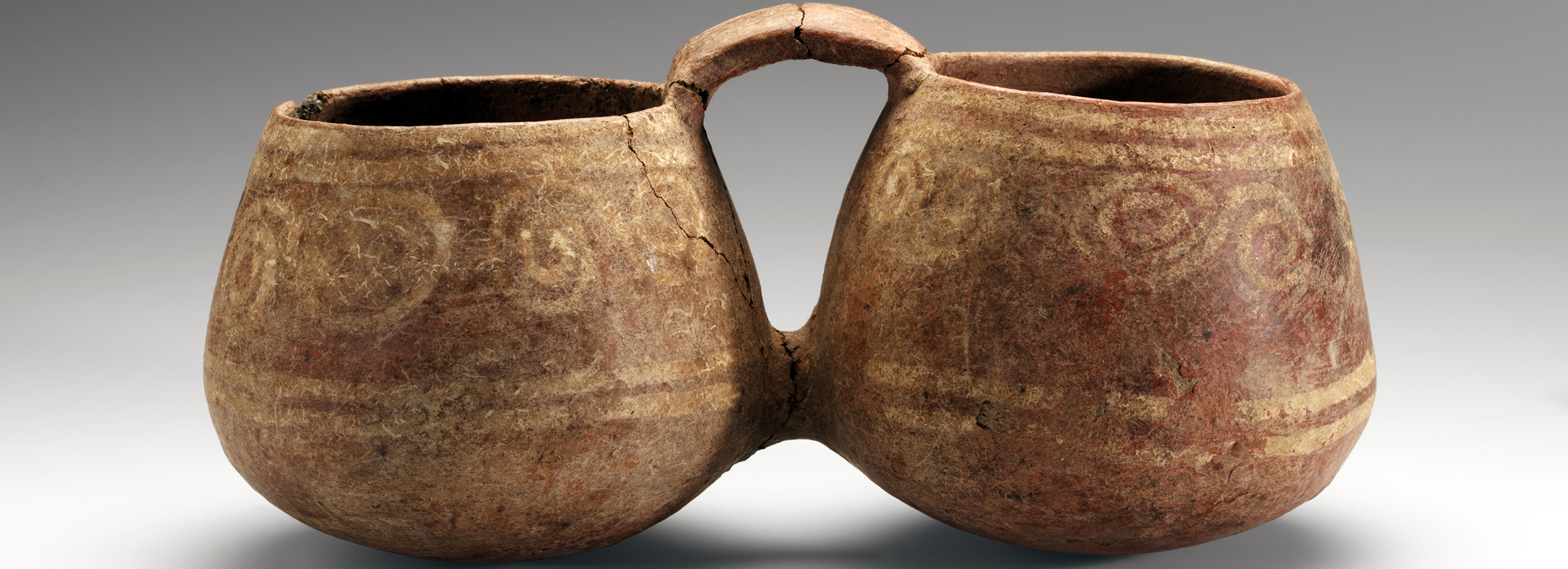 A light brown double-chambered ceramic vessel is painted with reddish stripes and swirls