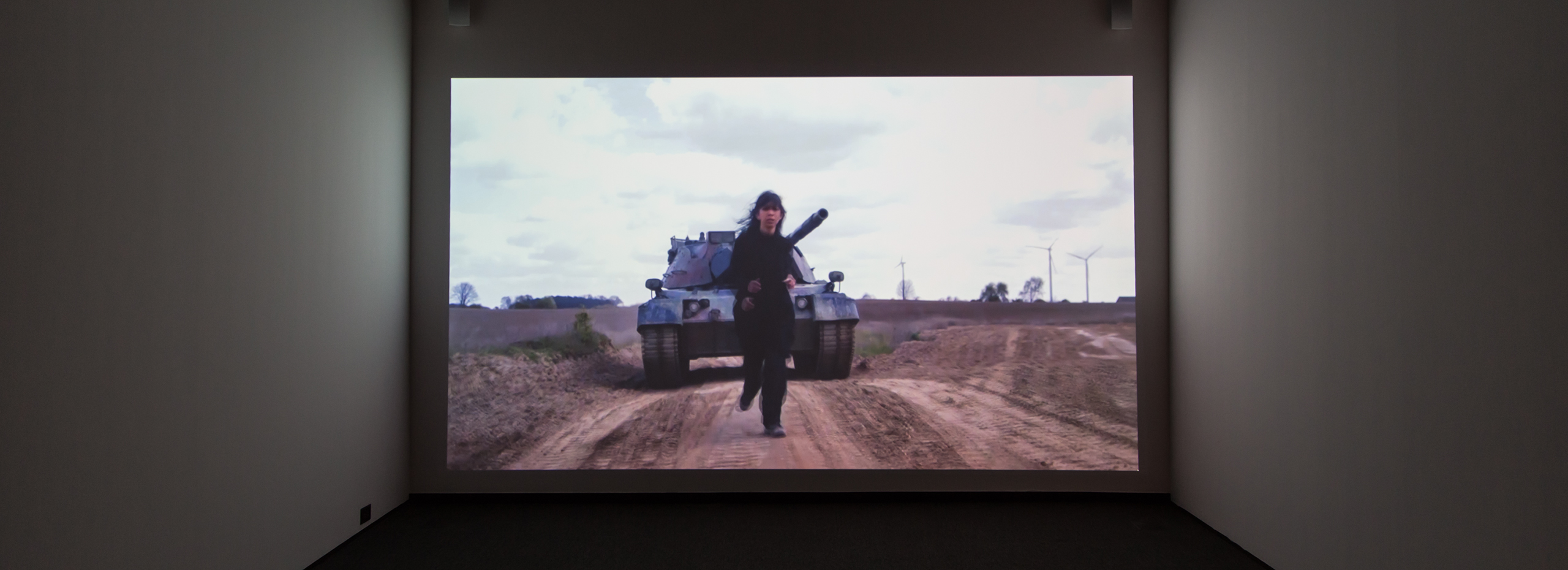 Projected film shows a dark-haired woman in dark clothing running away from a military tank down a dirt road.