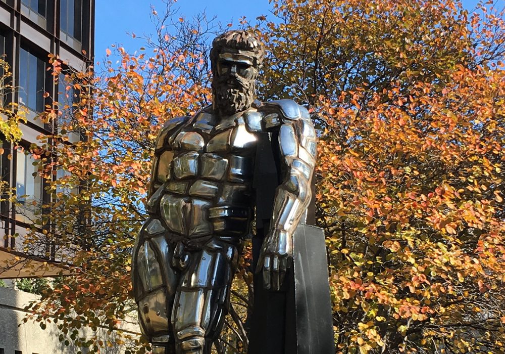 A large statue of a naked man made of chrome is installed between buildings and surrounded by autumn trees