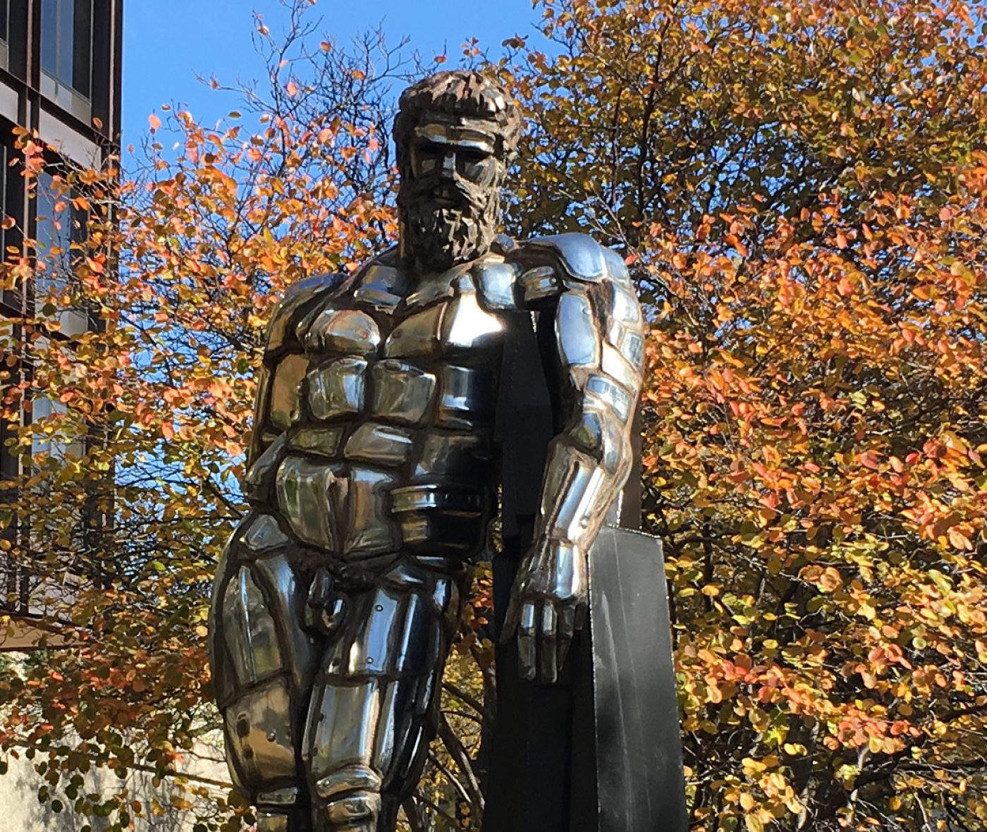A large statue of a naked man made of chrome is installed between buildings and surrounded by autumn trees