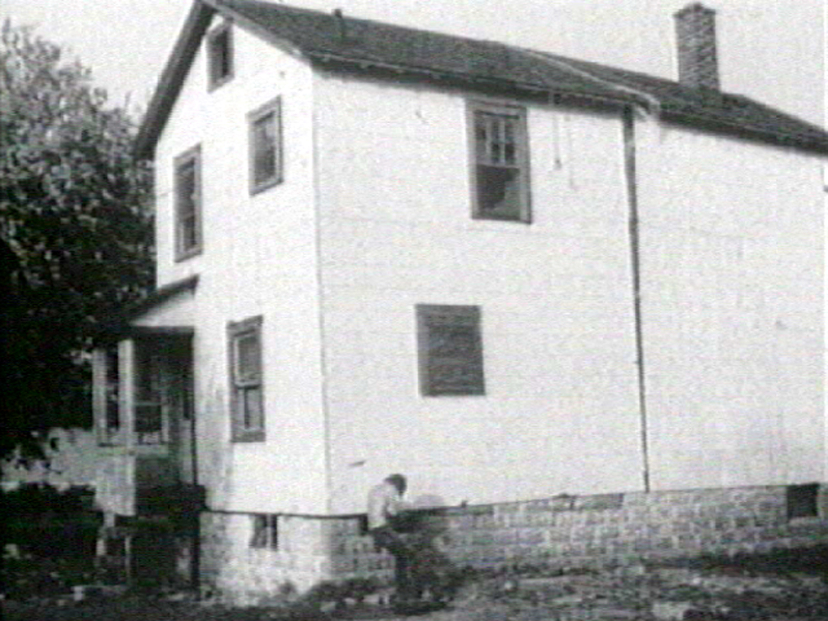A black and white image of a two-story white house with a man standing near its foundation line