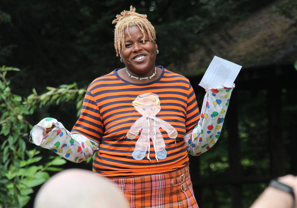 A Black woman with blonde hair wearing colorful clothes addresses an outdoor crowd