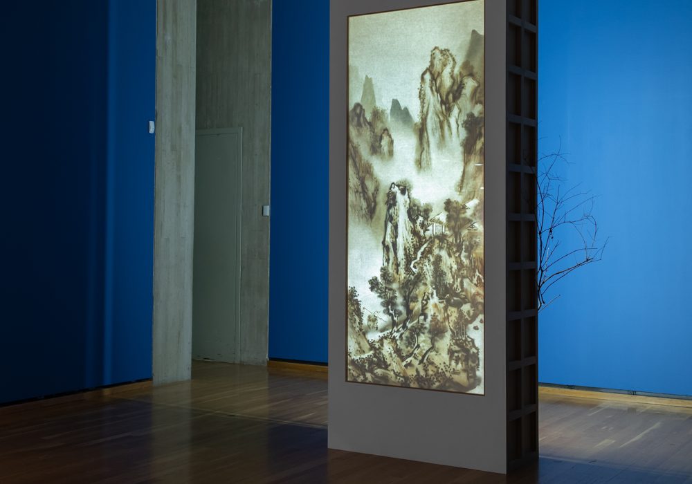 A tall lightbox with a branch sticking out projects an illuminated Chinese scroll in the center of a dim gallery with blue walls and wood floors