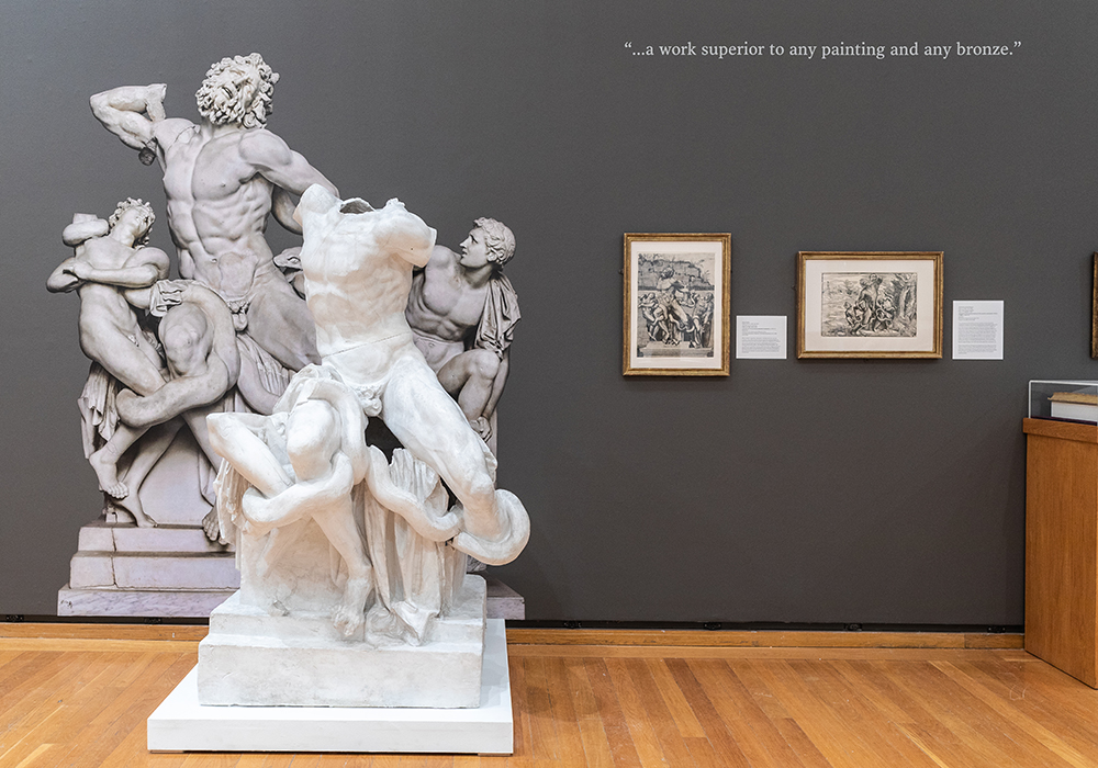 A plaster cast in a museum gallery is displayed in front of a photo of the original sculpture
