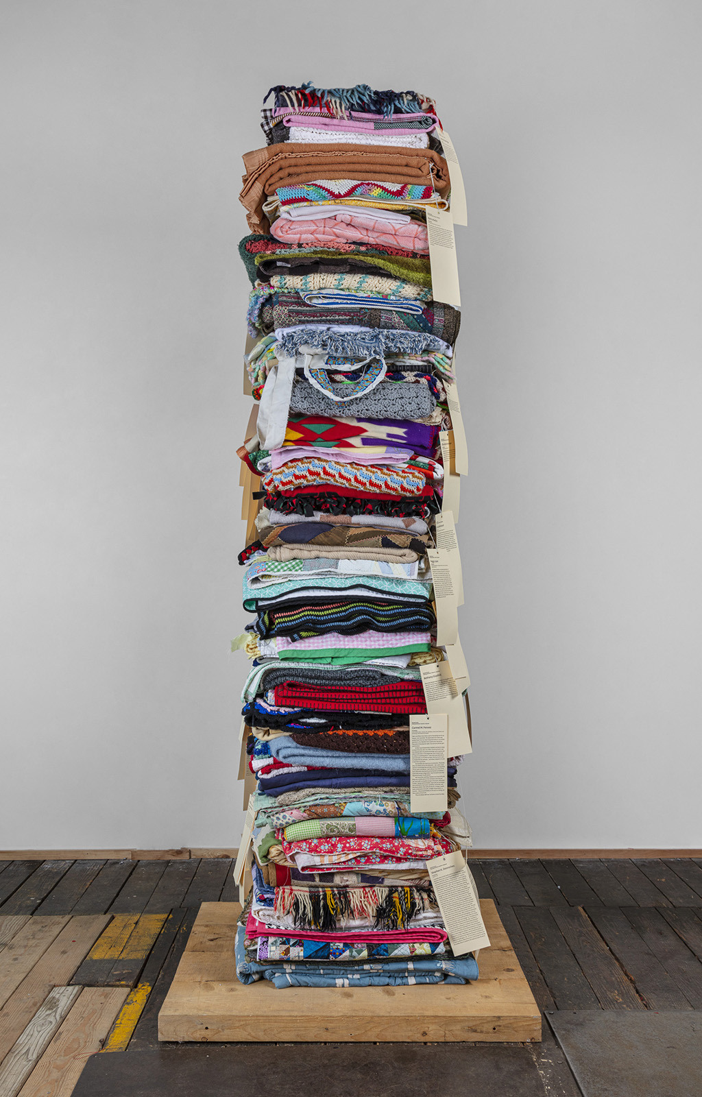 A tower of folded blankets in many colorful patterns is placed on a wooded base and long paper labels with text hang off the individual blankets