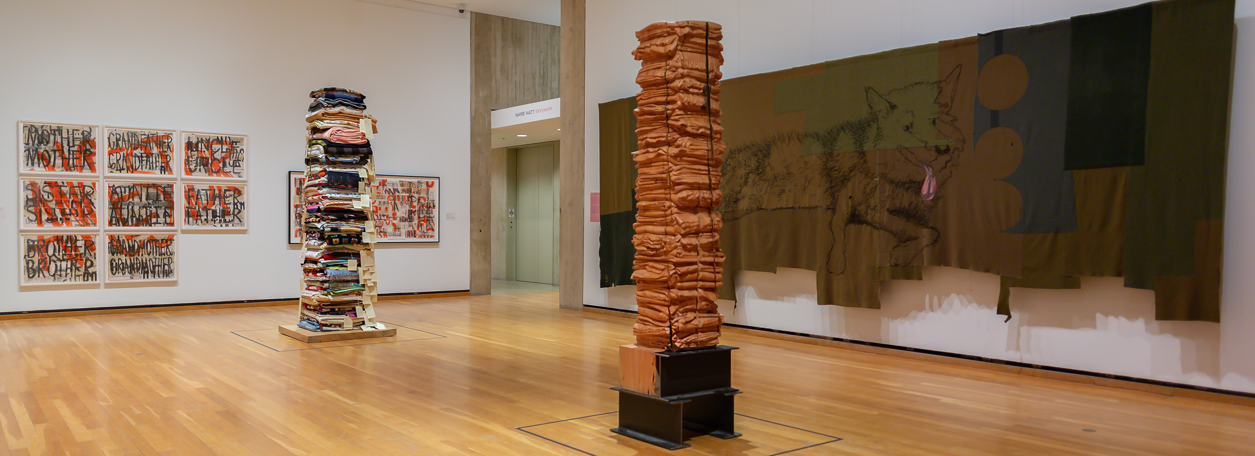 A museum gallery with two tall pillar sculptures in the center, framed prints hanging on one wall, and a large dark textile hanging on the other wall