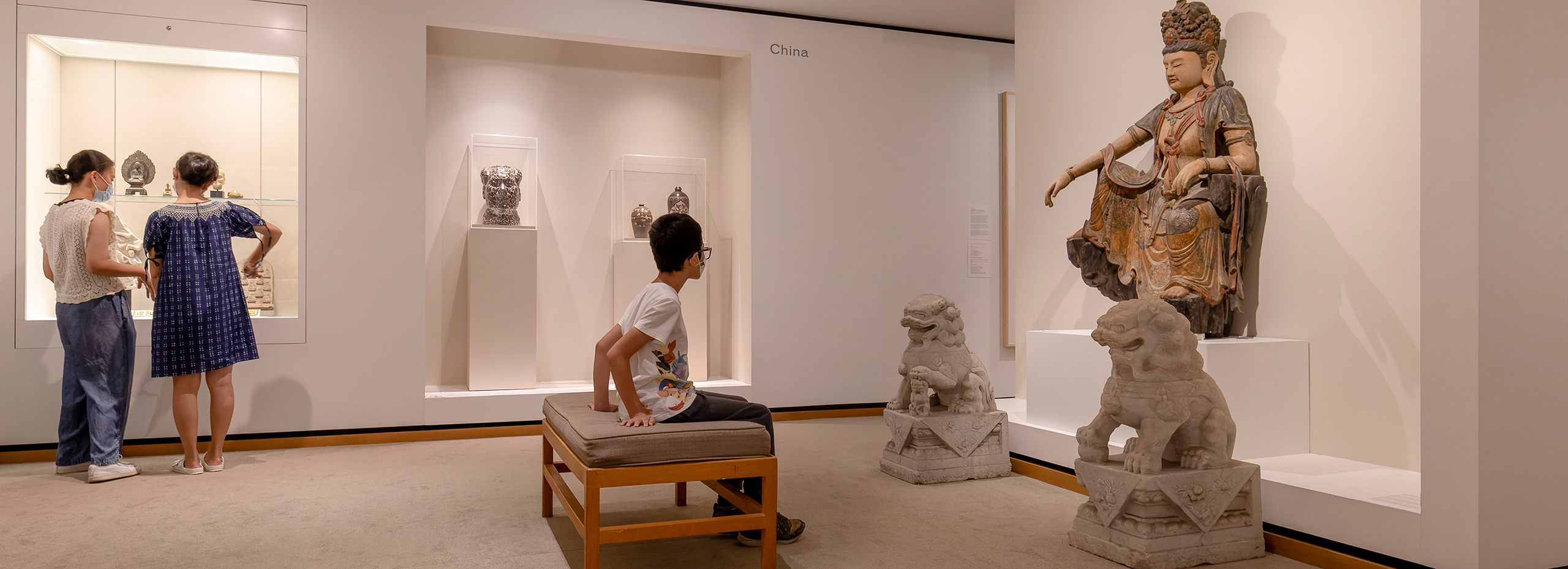 Two women and a boy visit a gallery of art from China including a large statue of a goddess flanked by stone lions