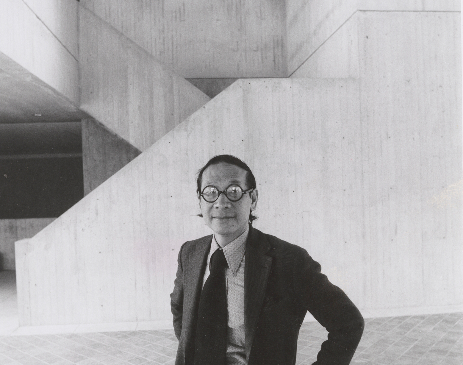 A black and white photo of Chinese American architect Ieoh Ming (I.M.) Pei standing in the lobby he designed of the Herbert F. Johnson Museum of Art, which has tall, angular concrete forms.