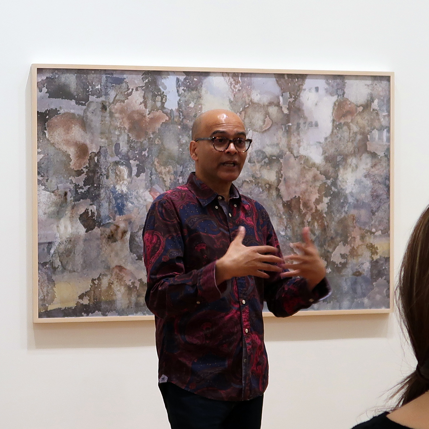 A bald man in glasses talks and gestures in front of a framed artwork on the wall behind him