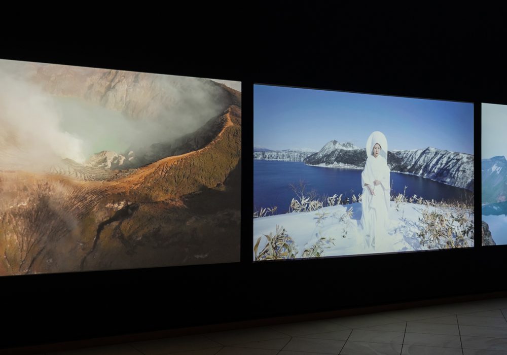 A three-channel video projection shows a central woman in white in and beside landscapes of clouds, mountains, and bodies of water