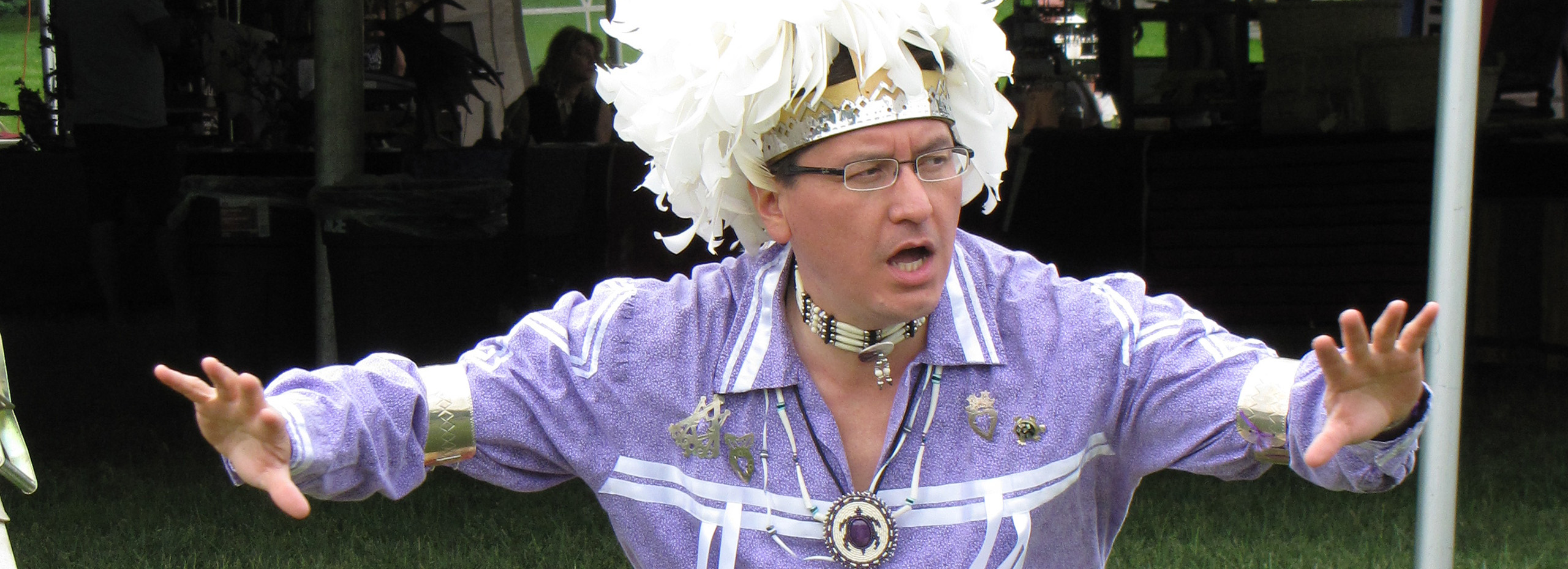 A man wears a white headdress, glasses, beaded necklaces, silver jewelry, and a purple shirt with white stripes while he opens his arms wide in the middle of telling a story