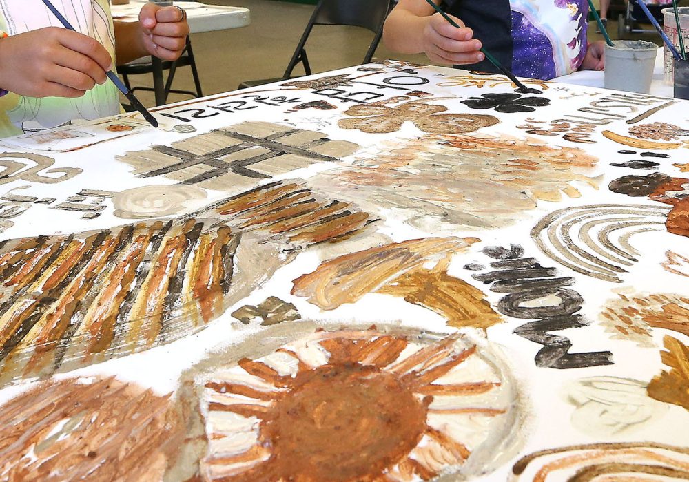 The hands of two children are seen working on a tabletop painting of different shapes in browns and blacks
