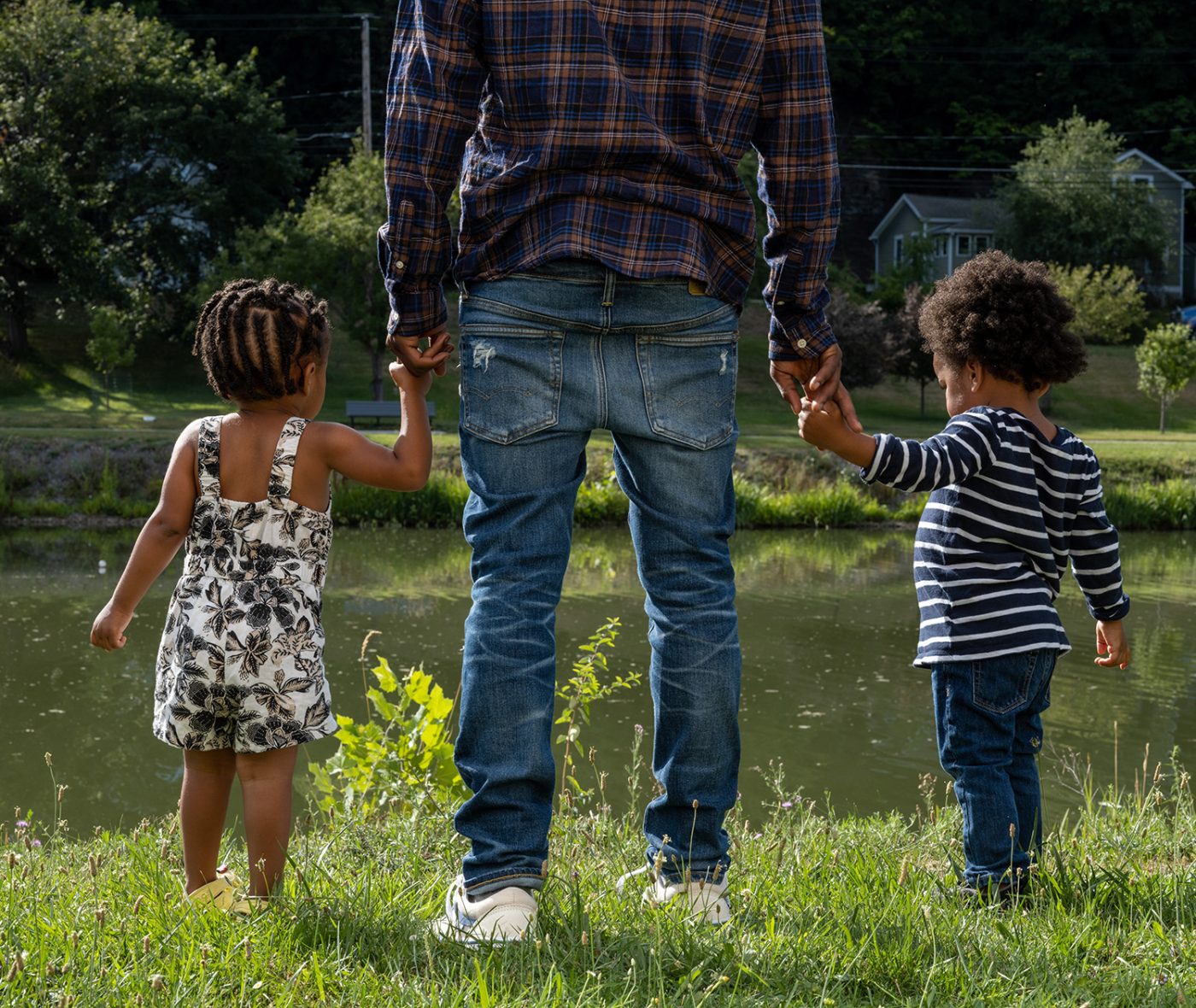 A Black man holds the hands of a Black boy and a Black girl next to a pond on a summer day