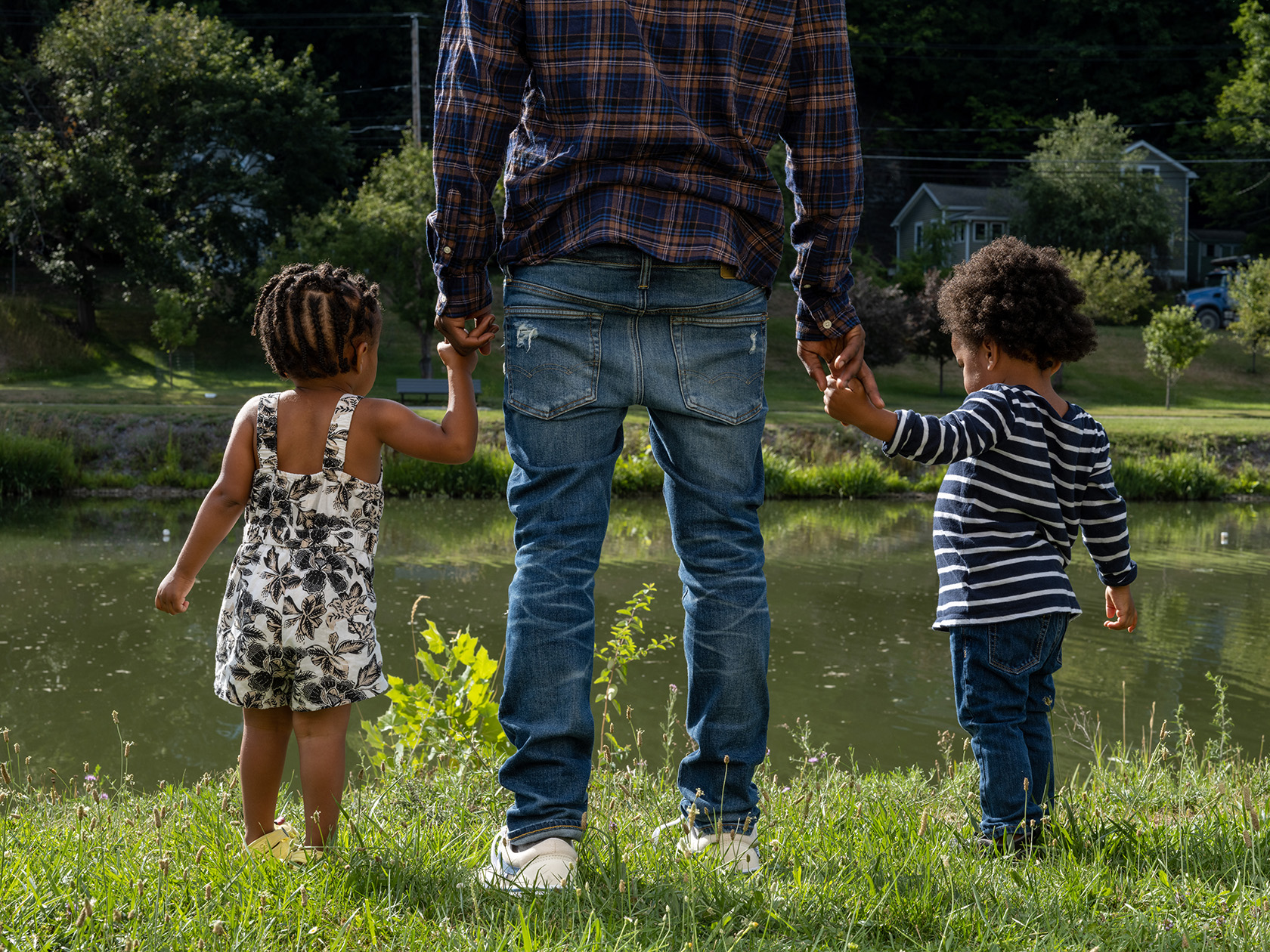 A Black man holds the hands of a Black boy and a Black girl next to a pond on a summer day