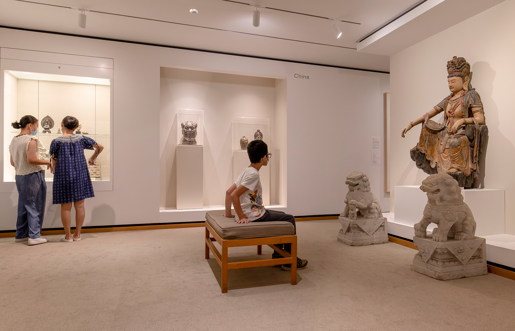 Two women and a boy visit a gallery of art from China including a large statue of a goddess flanked by stone lions