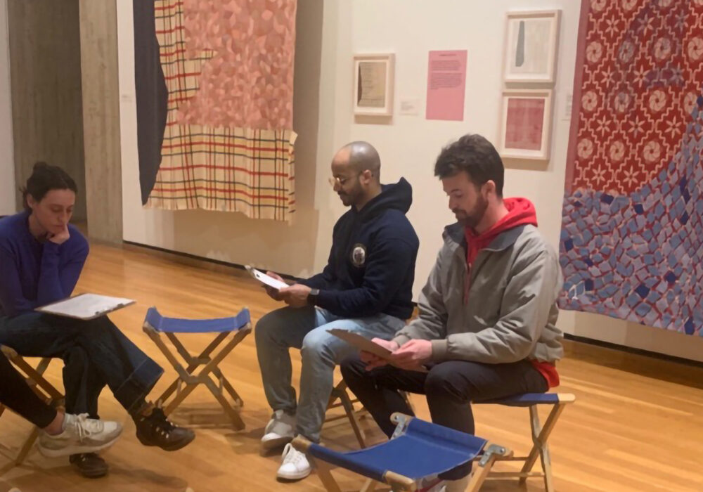 A group sits in an art gallery with colorful textiles hanging on the white walls