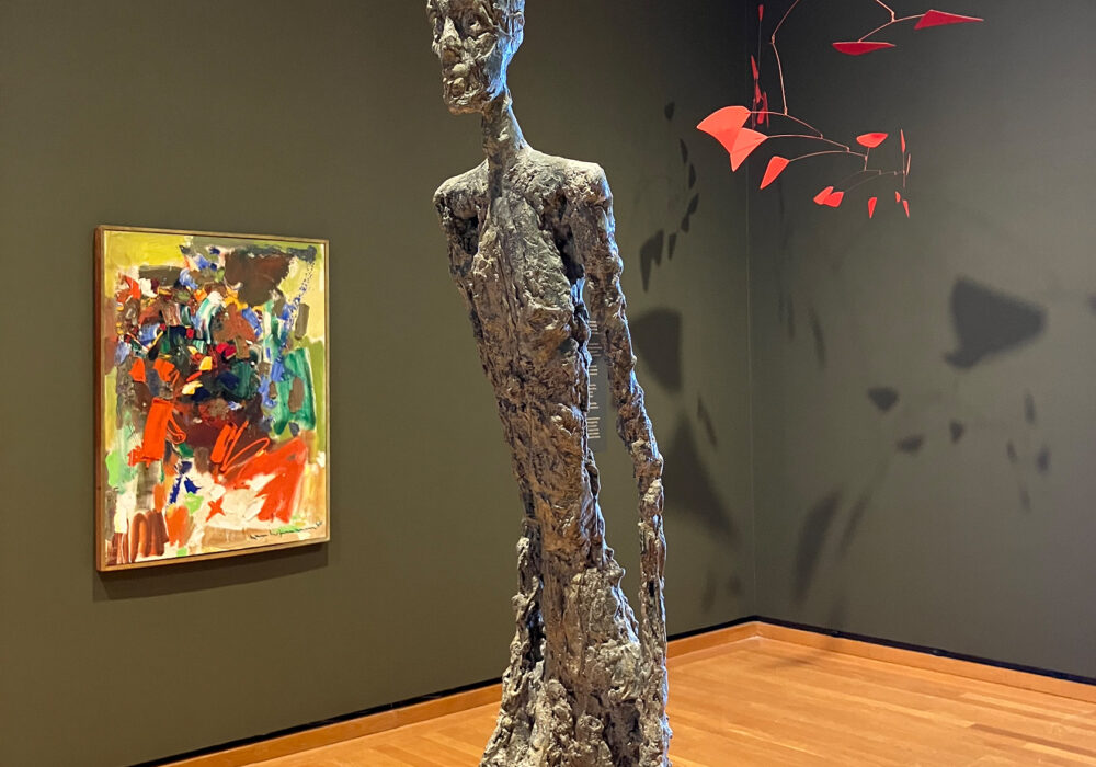 A colorful painting, thin figural sculpture, and red mobile in a museum gallery with dark green walls