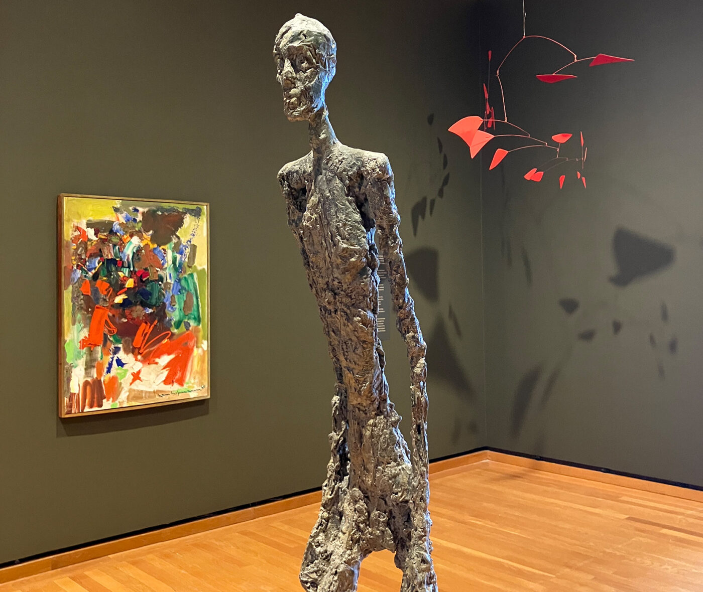 A colorful painting, thin figural sculpture, and red mobile in a museum gallery with dark green walls