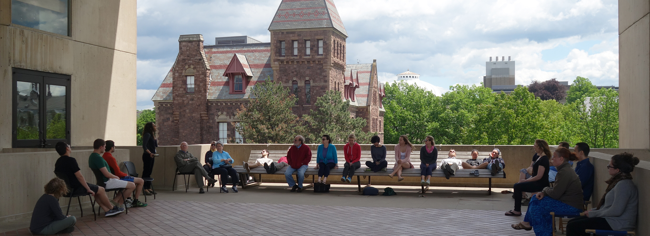 People sit on a long bench and chairs in an outdoor space with buildings in the background
