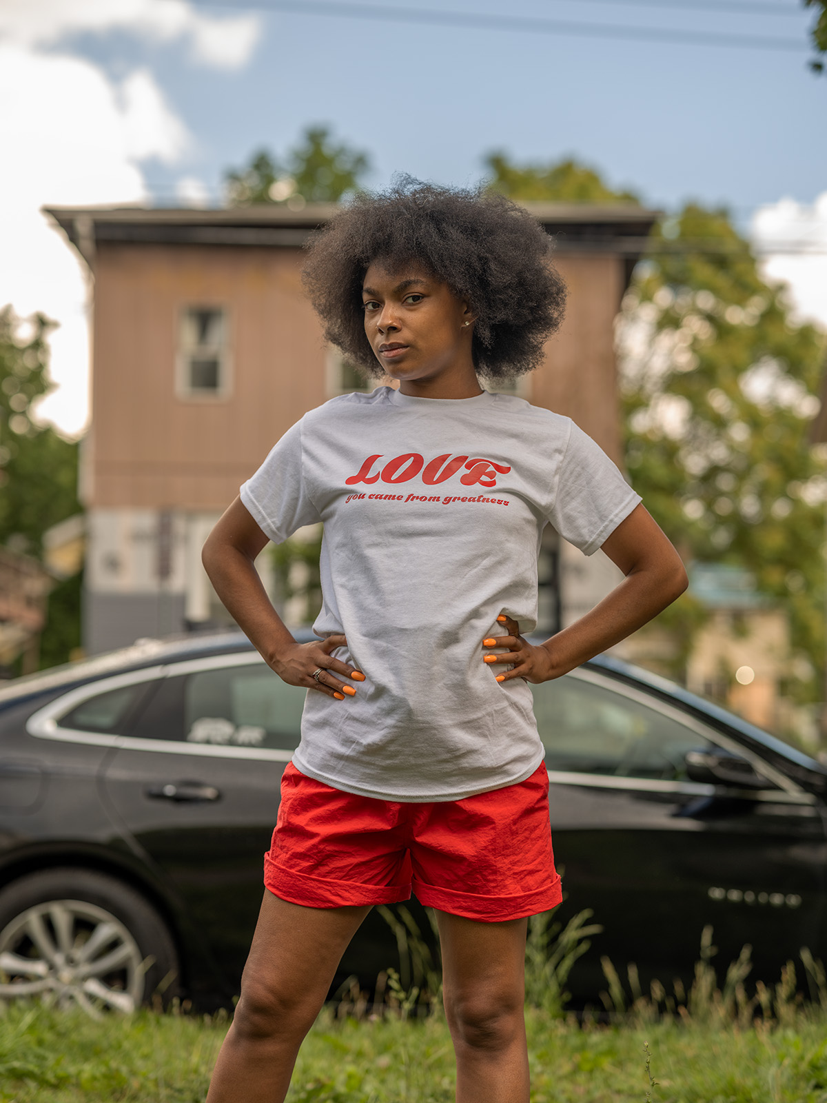 A young Black woman in shorts and a T-shirt stands outside in front of a black car