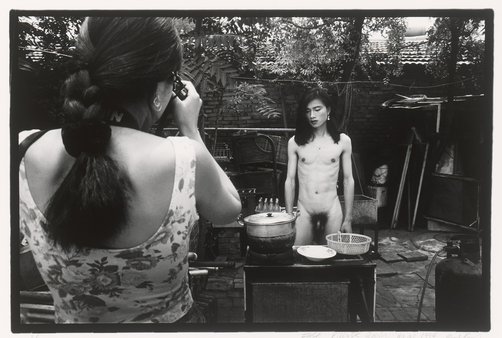 A black and white photograph showing a woman taking a photo of a naked man with long hair