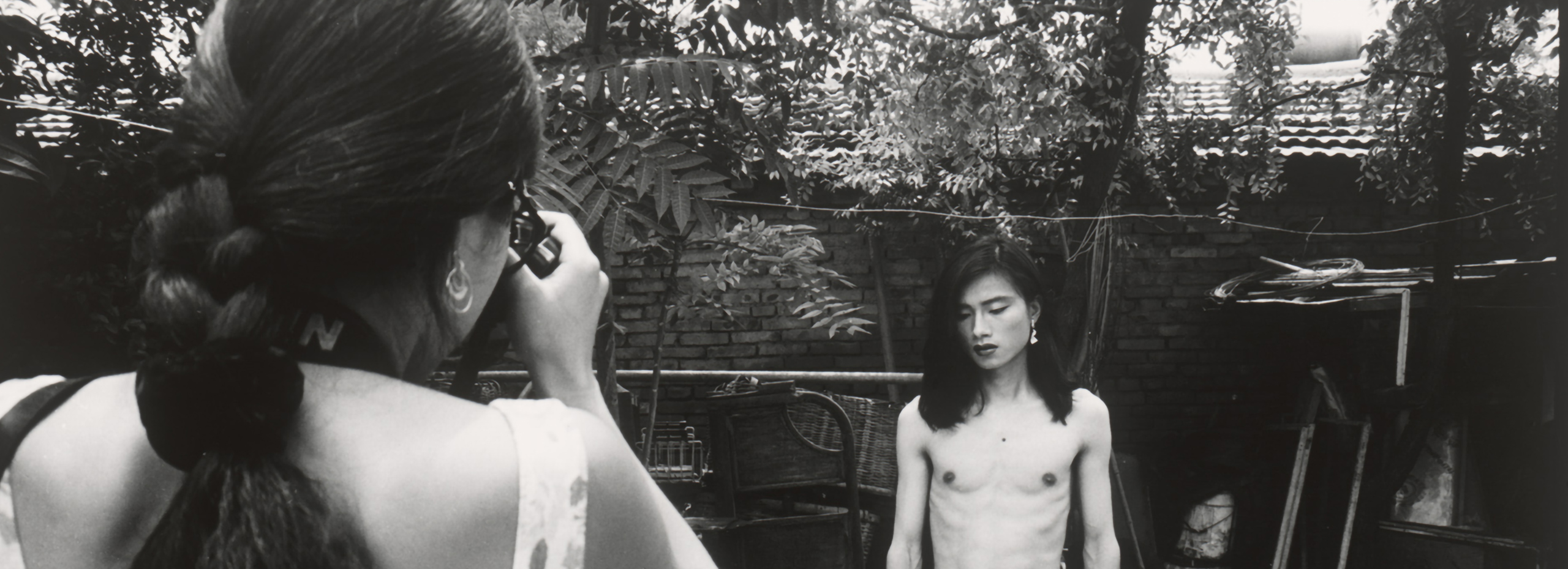 A black and white photograph showing a woman taking a photo of a shirtless man with long hair