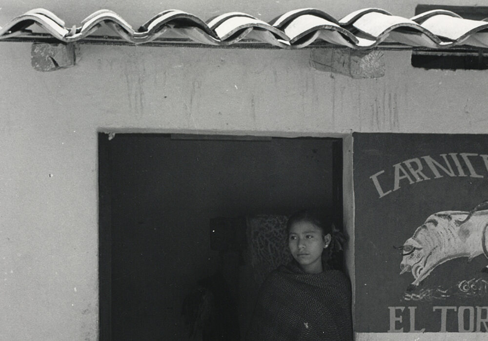 A black and white photograph of a young woman standing outside a butcher's market in Mexico