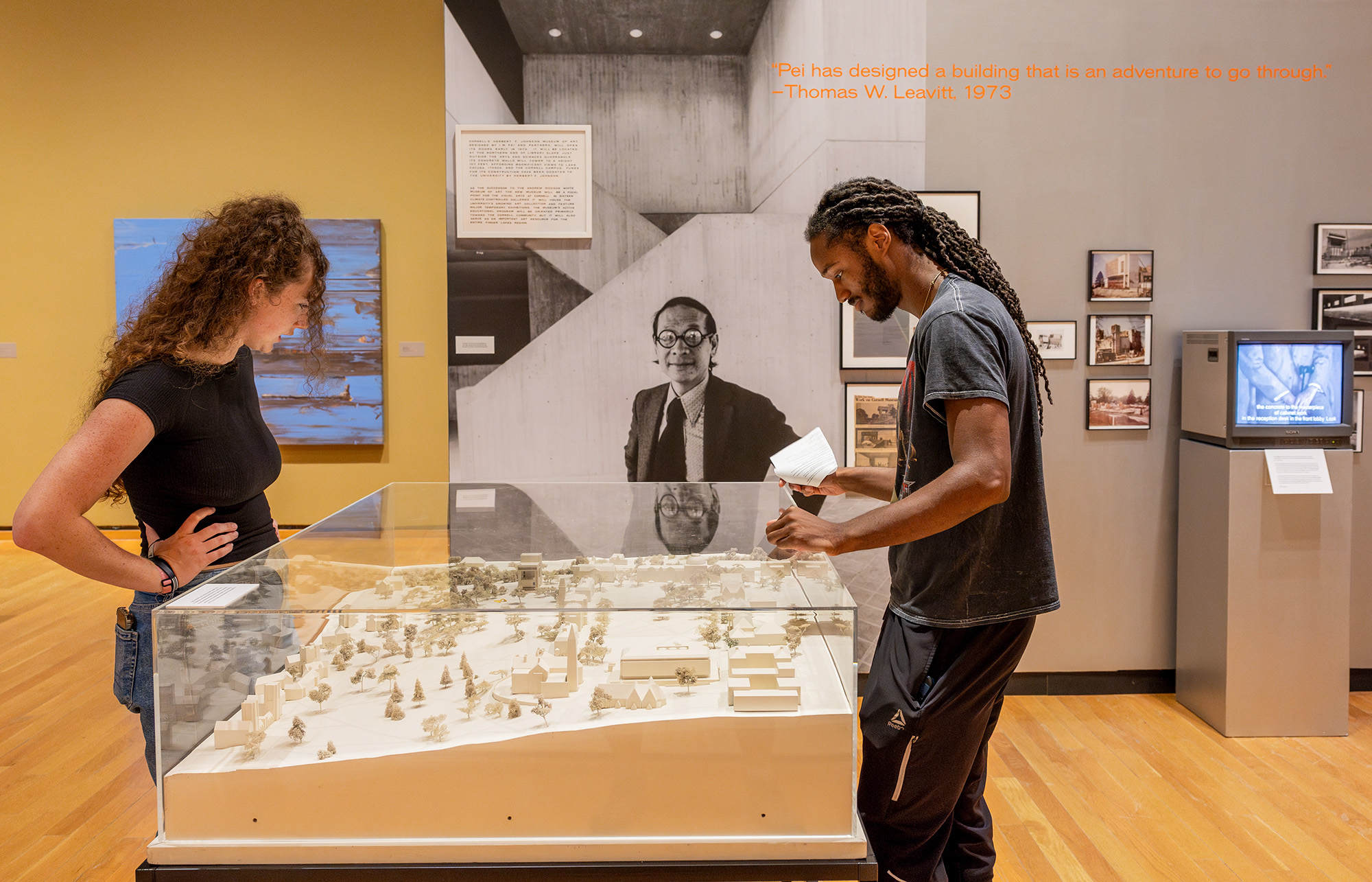 A man and woman look at a white architectural model in a museum gallery with art and a large photo of architect IM Pei