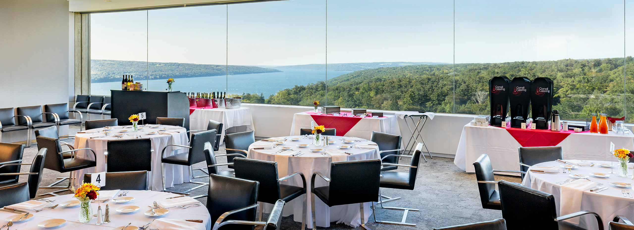 Round tables are set for a meal with black chairs in a large room with a lake view