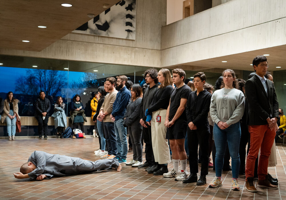 A group of people stand in a circle as an artist wearing a gray suit lies on a tile floor in a performance at the Johnson Museum of Art