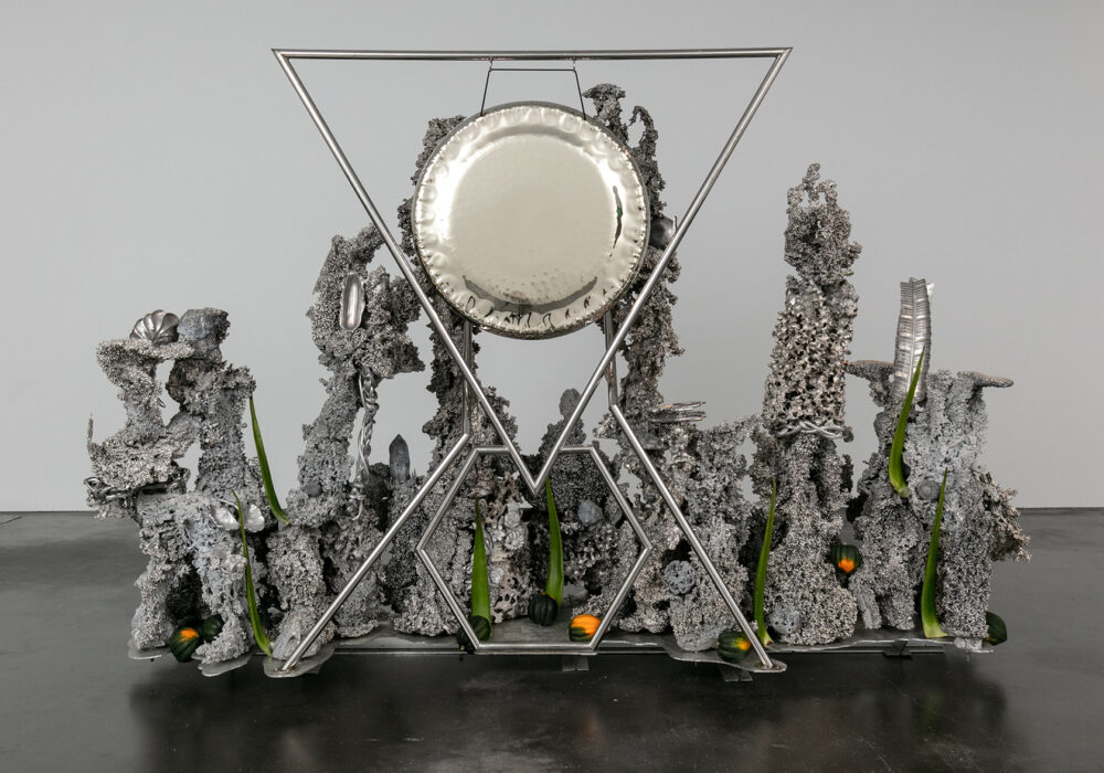 A large silver sculpture with organic forms and geometric lines framing a silver gong
