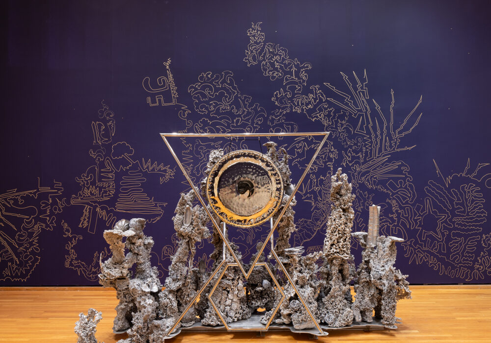 A large silver sculpture with organic forms and geometric lines stands against a dark purple wall with an elaborate line drawing and wave pattern