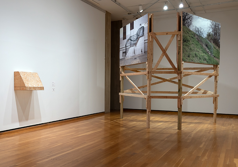 Two sculptures, one resembling plywood in the shape of an oven range hood on a wall and the other a billboard structure with two large images of outdoor stairs