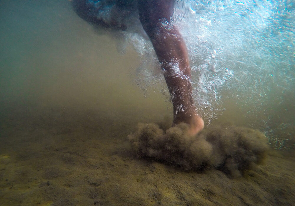 Detail of a photograph showing a Black leg stepping into underwater sand