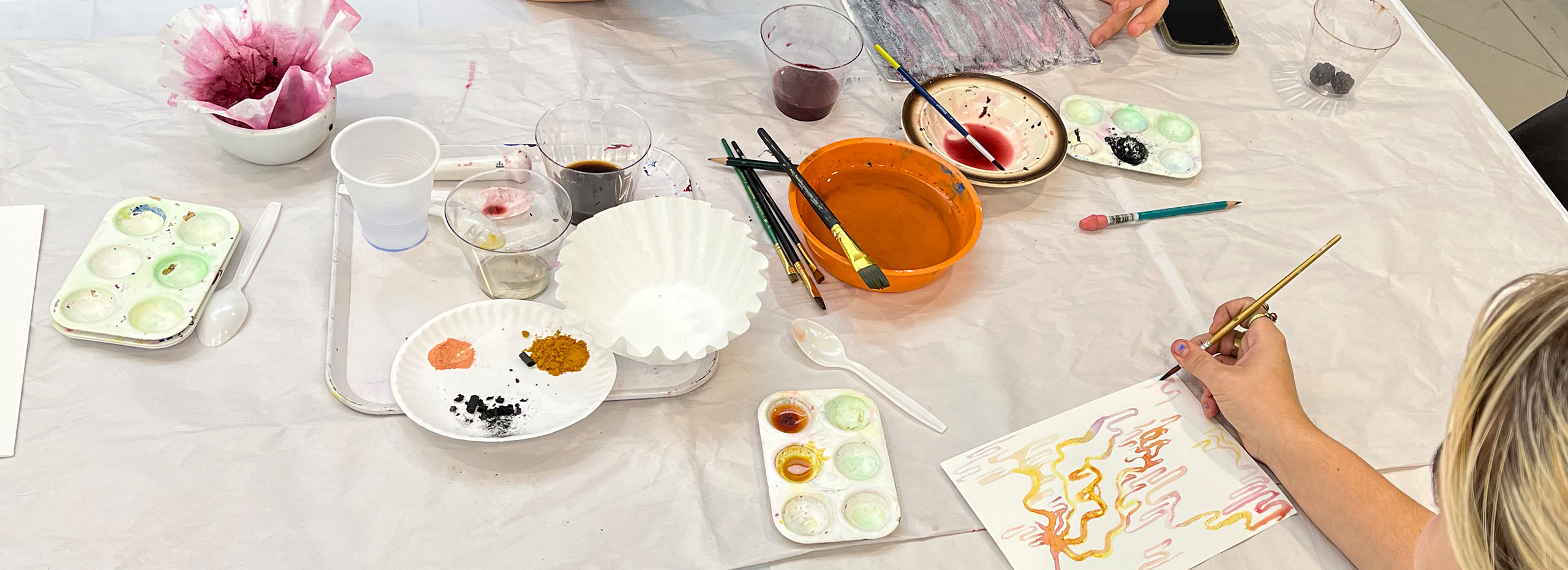 Hands use watercolor painting supplies on a table with a white tablecloth