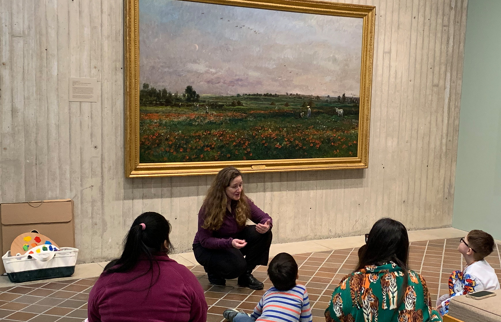 A teacher sits on a floor in front of a large painting and talks to schoolchildren