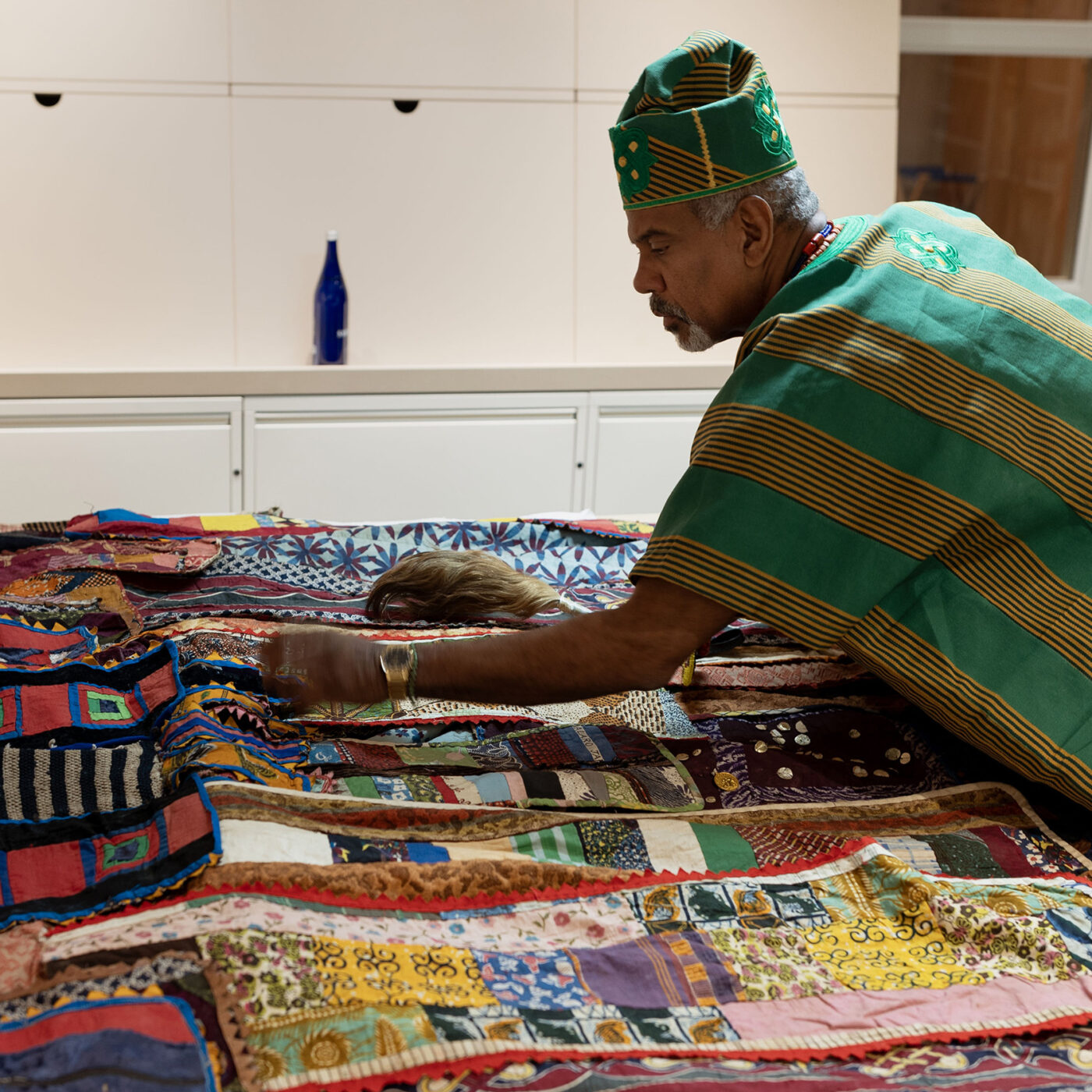 A Yorùbá priest in formal dress touches a large colorful textile on a table