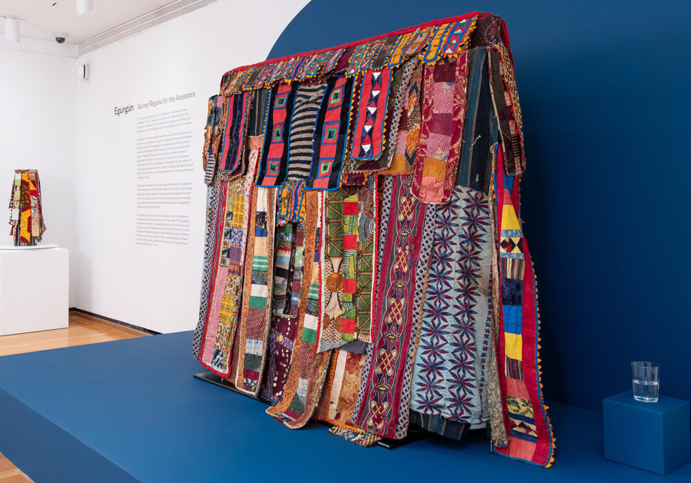 A colorful West African regalia is displayed on a dark blue platform in a museum gallery