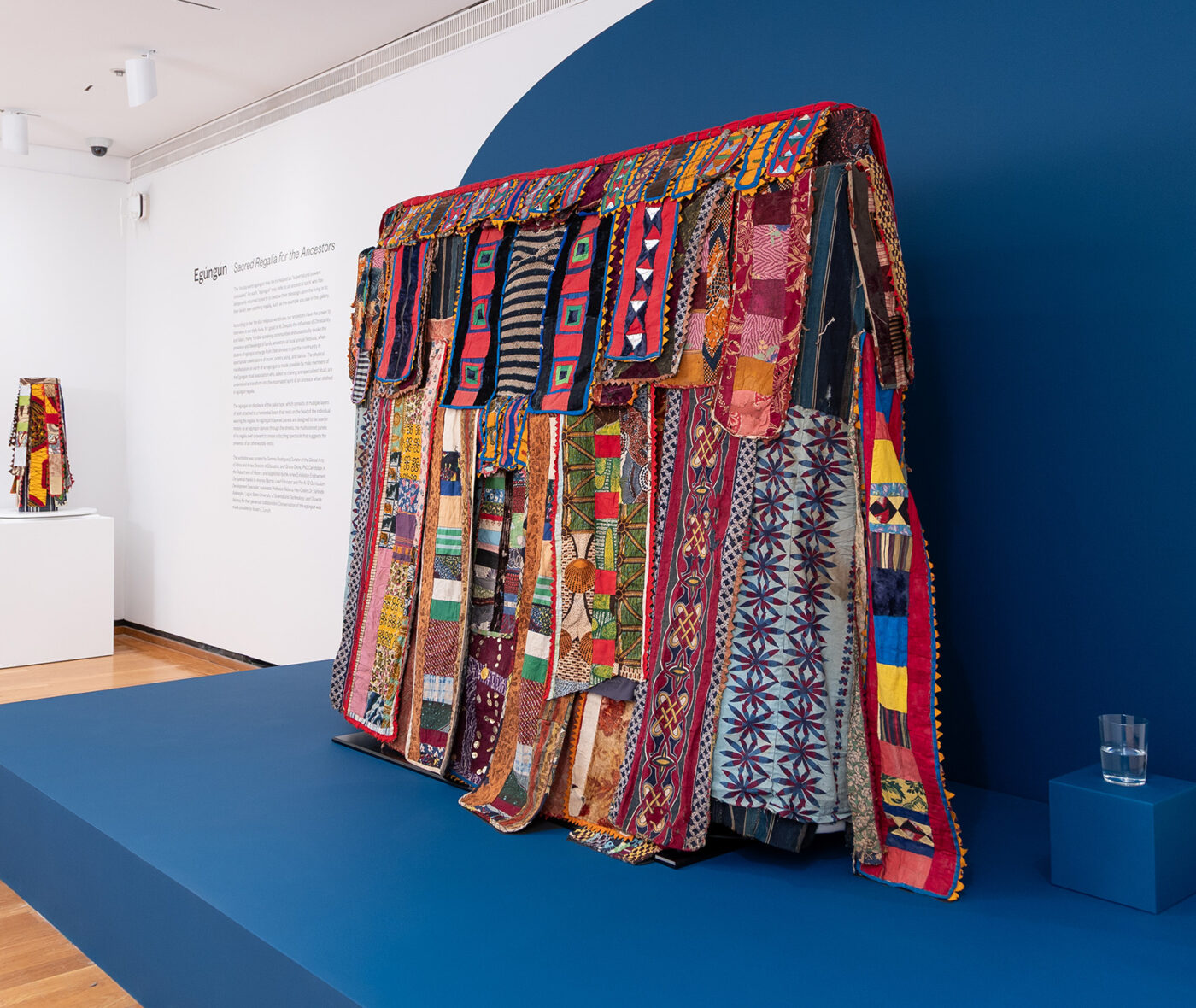 A colorful West African regalia is displayed on a dark blue platform in a museum gallery