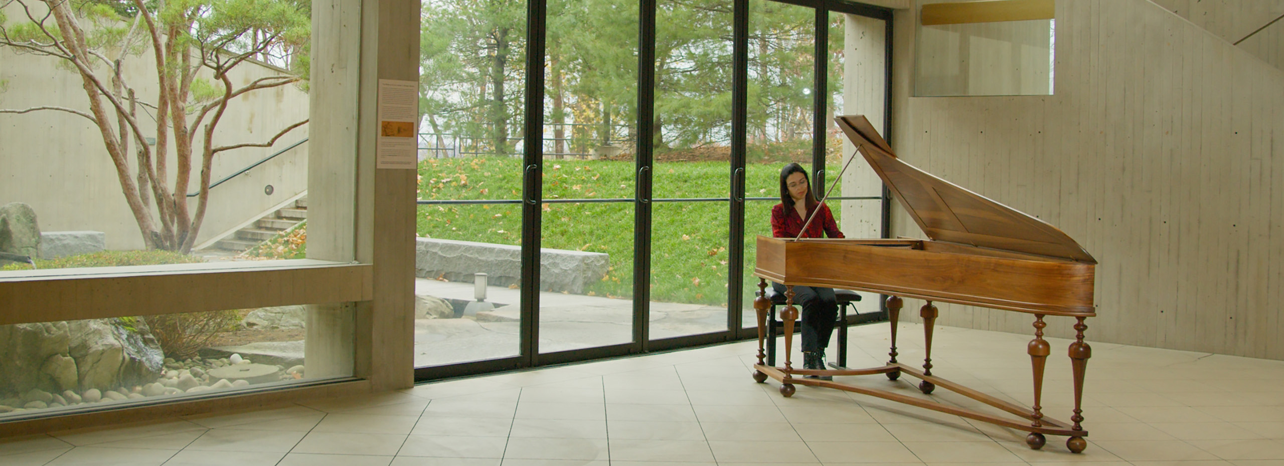 A woman plays the piano next to large windows leading to a garden outside