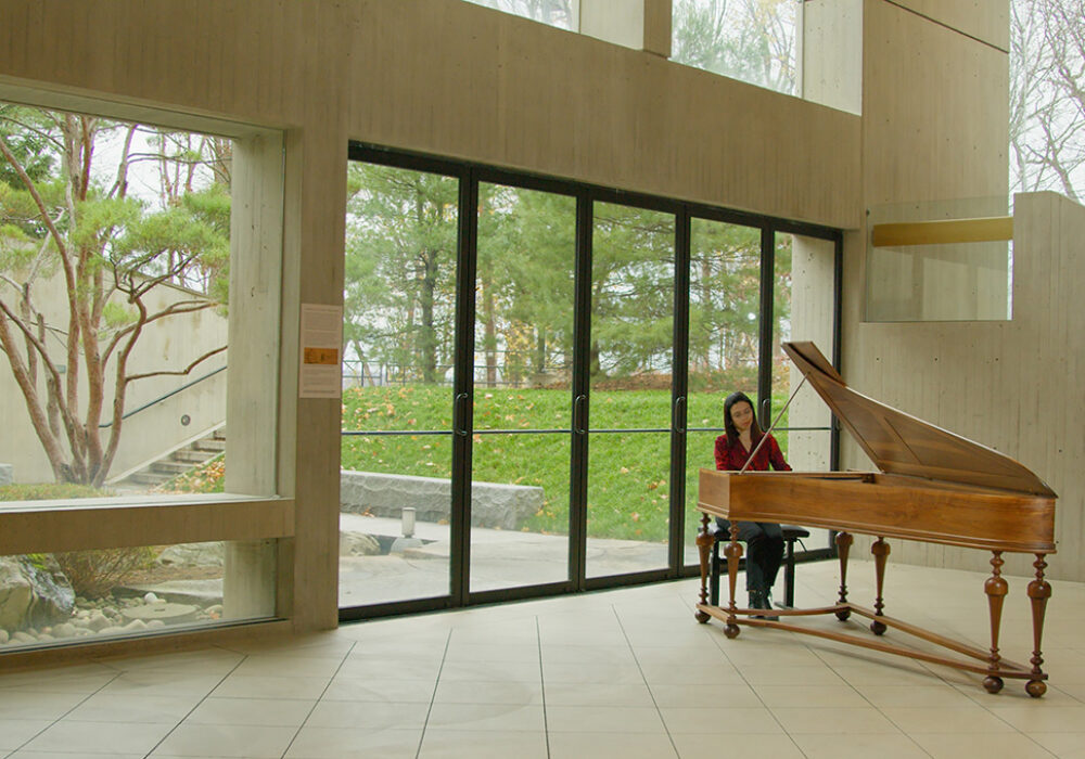 A woman plays the piano next to large windows leading to a garden outside