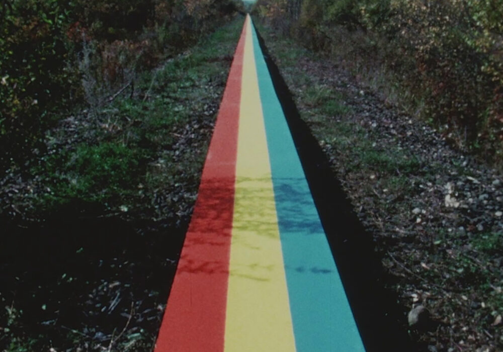 Video still of a long plank painting with red, yellow, blue, and black stripes extends into the distance between rows of green trees
